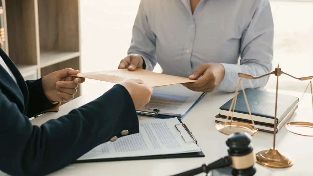 Two people exchanging an envelope over legal documents at a desk with scales of justice.